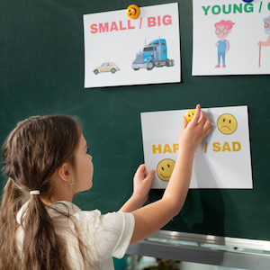 Dívka se učí anglicky ve škole u tabule, kde jsou obrázky s protiklady jako small big, young old a happy sad. Interaktivní a zábavná výuka angličtiny pro děti v jazykové škole TOPLINGVA.