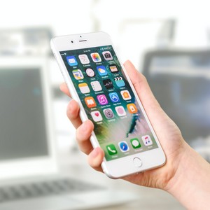 Ruka drží bílý chytrý telefon s aktivní obrazovkou s ikonami aplikací, v pozadí je rozmazaný interiér kanceláře.