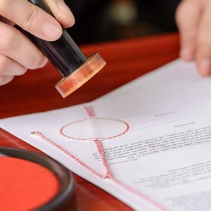 Žena, která aplikuje kulaté razítko na svázaný úřední dokument s červeným provázkem a tiskovou podložkou.