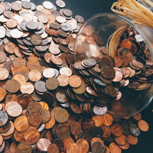 Skleněná nádoba převrácená na tmavém povrchu, z níž se sypou a jsou rozházeny měděné mince.