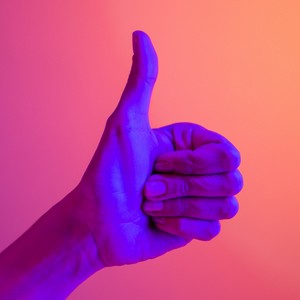 Ruka se zdviženým palcem (znamení souhlasu) osvětlená neonovým fialovo-oranžovým světlem.
