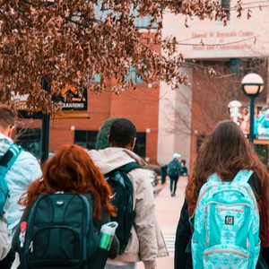 Studenti s batohy kráčející na univerzitní kampus se suchými podzimními listy na stromech.