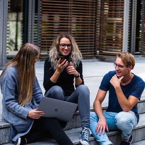 Tři mladí lidé sedí venku na šedých schodech: dvě ženy a jeden muž, žena uprostřed ukazuje na svůj mobilní telefon a usmívá se.