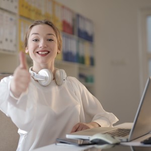 Usmívající se mladá žena sedí u stolu s notebookem a zdviženým palcem, na krku má bílá sluchátka.