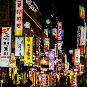 Večerní pohled na rušnou ulici plnou svislých a vodorovných, zářivě osvětlených korejských neonových reklam a nápisů.