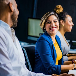 Žena v modrém saku sedí u stolu s kolegy a usmívá se do fotoaparátu v moderní kanceláři.
